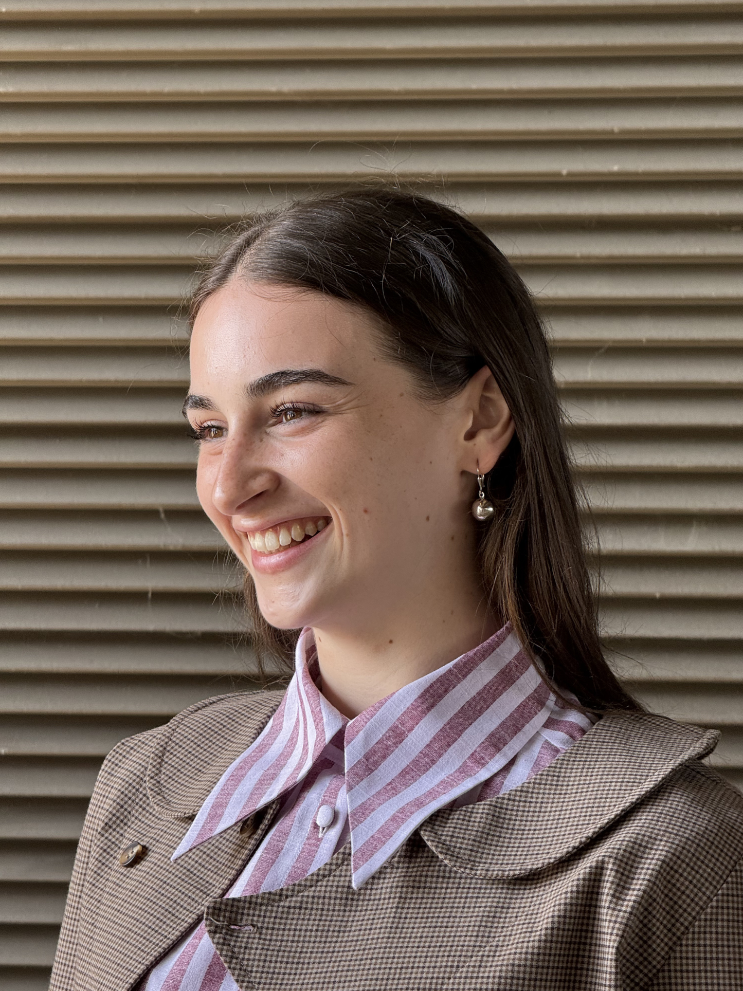 Woman wearing a stripe long collar shirt and brown coat against a corrugated metal background