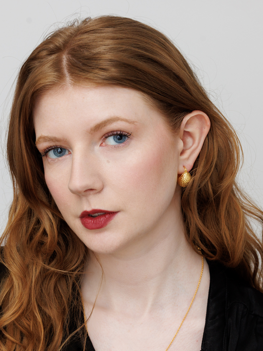 Woman with red hair and red lipstick wearing gold ball hoop earrings against a plain background