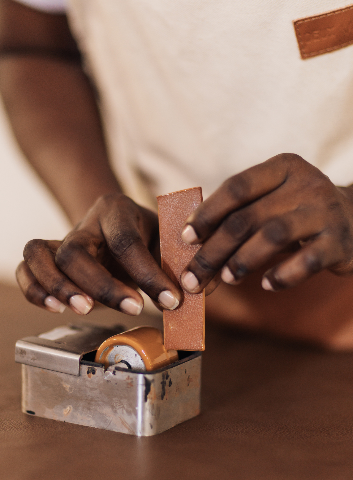 A close-up of a person's hands using a small metal leather roller tool on a tan leather strip, with a cream apron visible in the background.