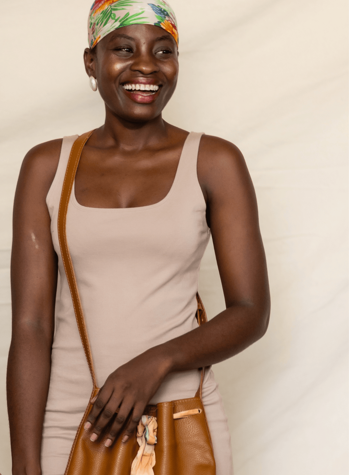 A smiling woman wearing a beige ribbed tank top and a colorful floral headwrap, carrying a brown leather shoulder bag against a light beige backdrop.