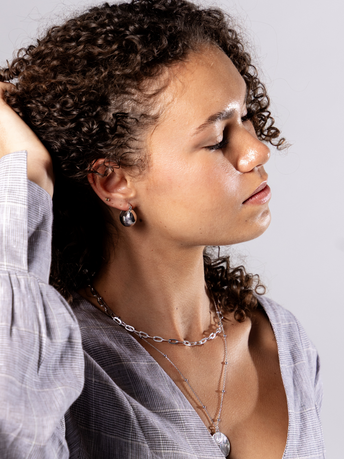 Woman with curly hair wearing a light grey dress and multiple silver necklaces and recycled silver ball hoop earrings against a plain background