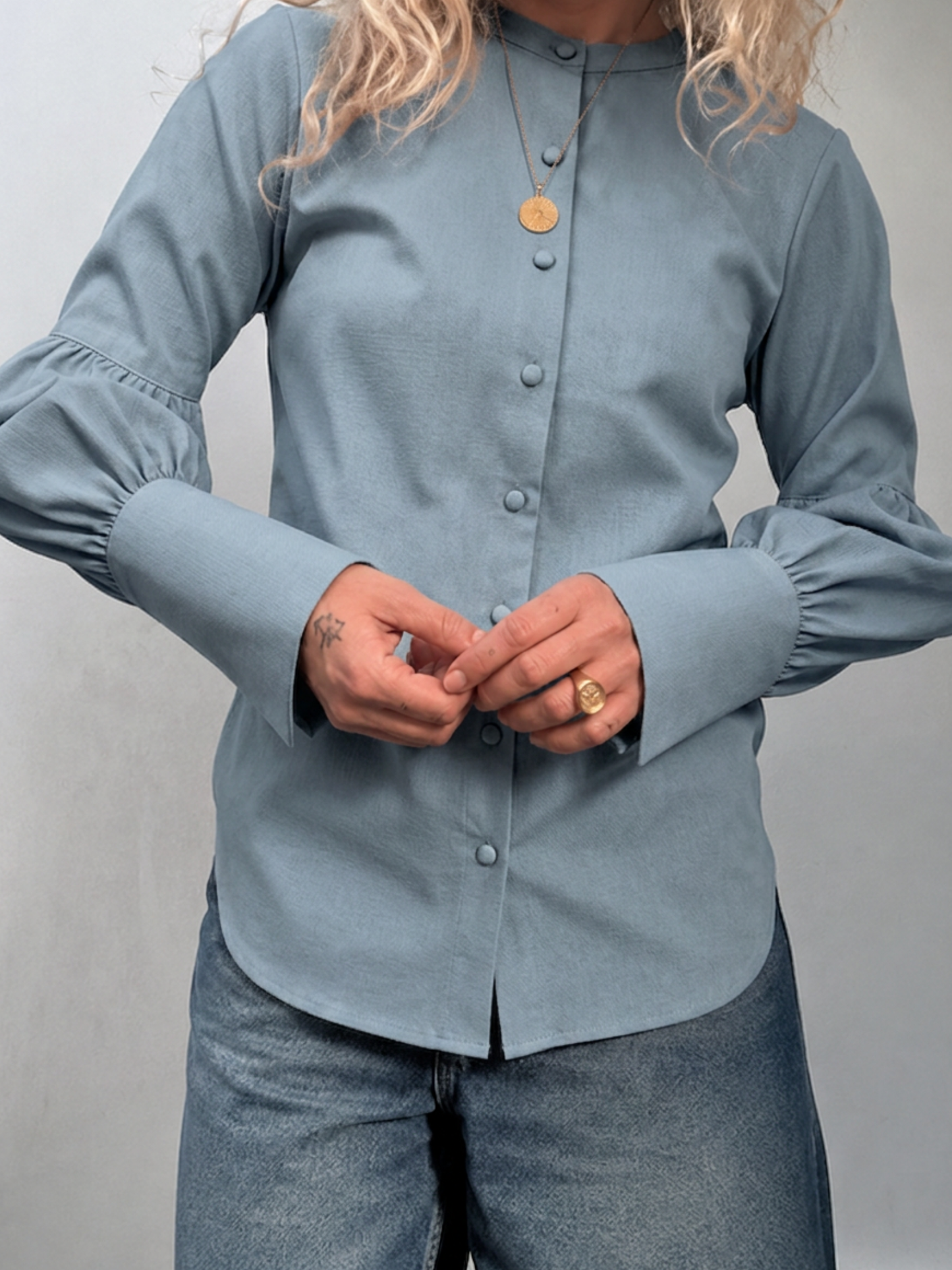 A woman wearing a dusty blue long-sleeve button-up blouse with gathered cuffs and blue jeans, shown from the shoulders down against a plain light gray background.