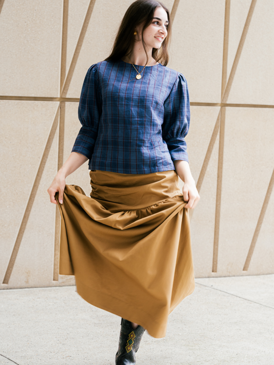 Woman wearing a blue plaid shirt and brown skirt against a geometric wall.