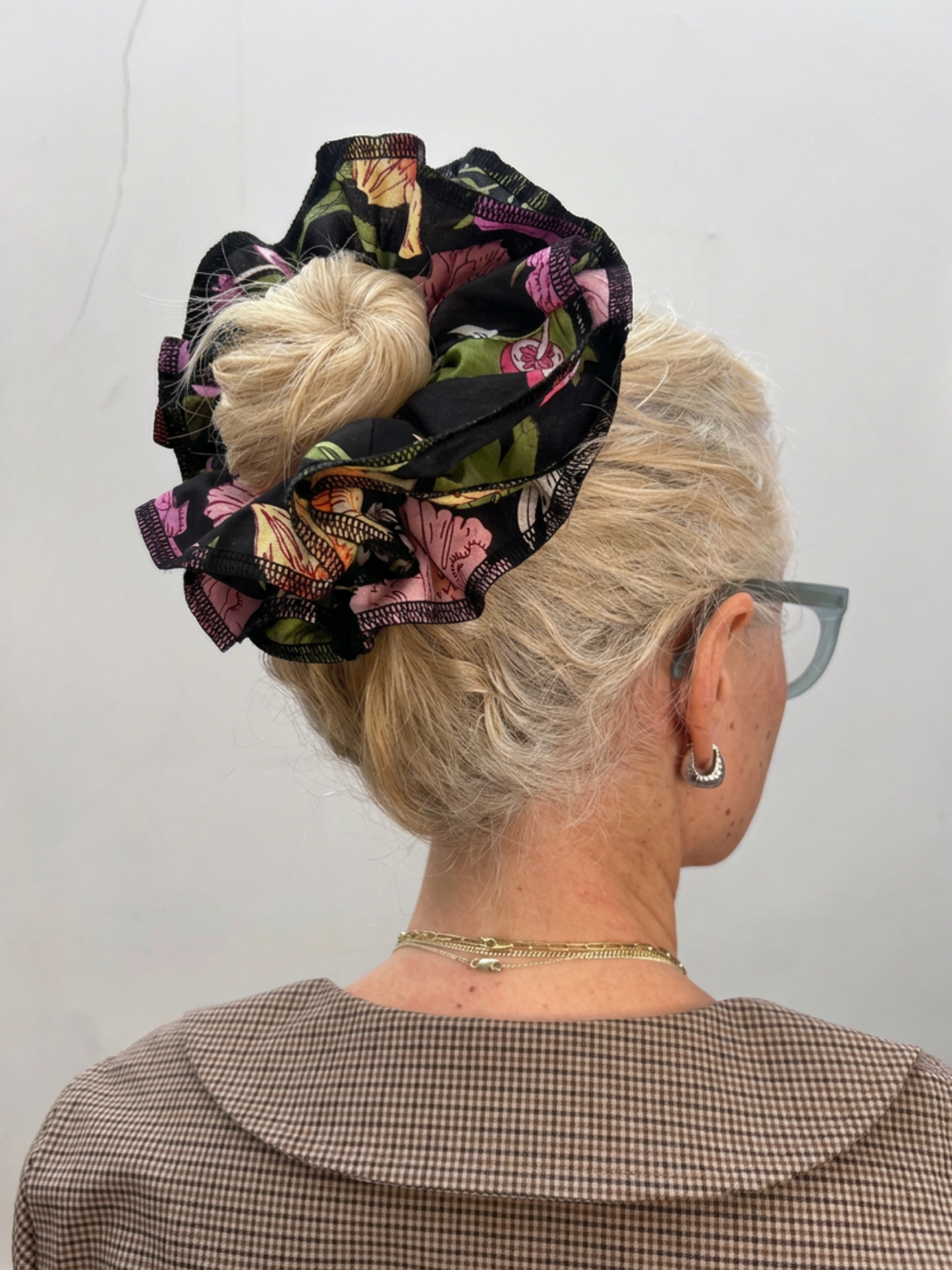 A blonde woman shown from the back wearing a large, ruffled hair scrunchie with a black, pink, and green floral pattern. She is also wearing glasses, a gold necklace, and a brown checkered top against a plain light gray wall.
