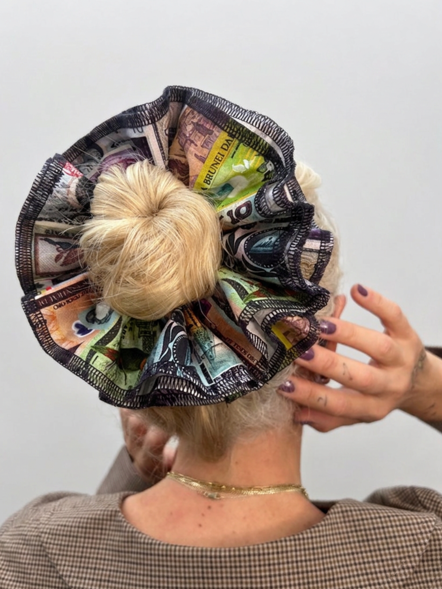 A large, ruffled hair accessory made from printed banknotes, styled in a bun on a person's head, shown from behind against a plain light gray background.
