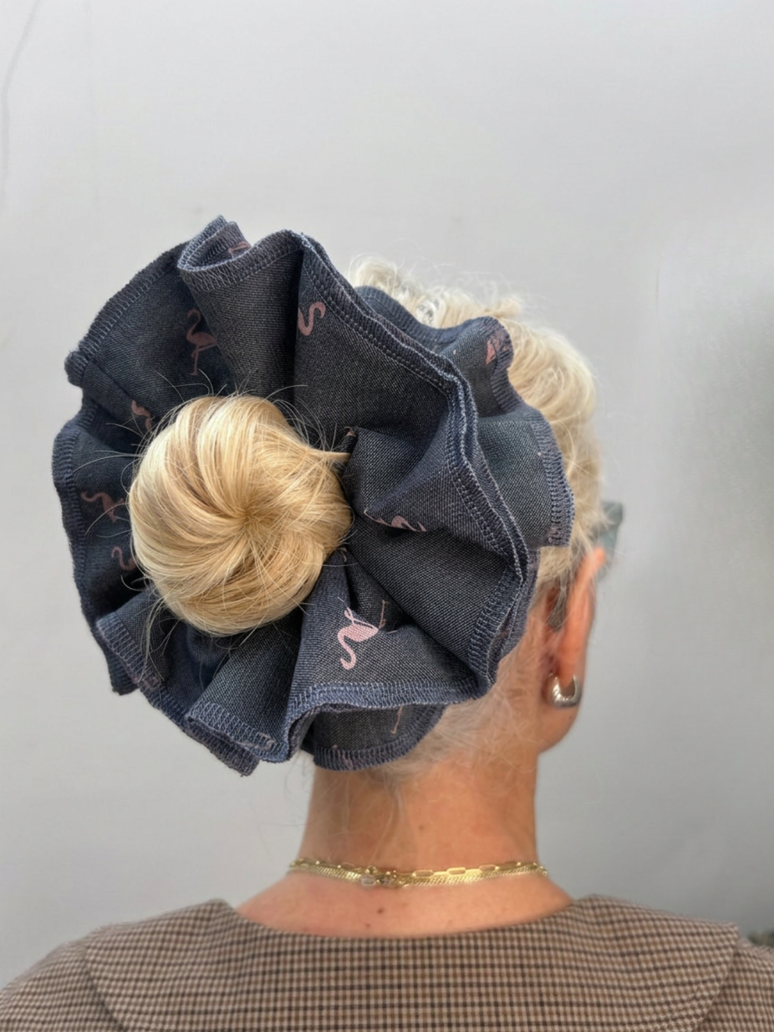 A blonde woman viewed from the back wearing a large fabric scrunchie in dark denim blue with pink flamingo prints, holding her hair in a bun.