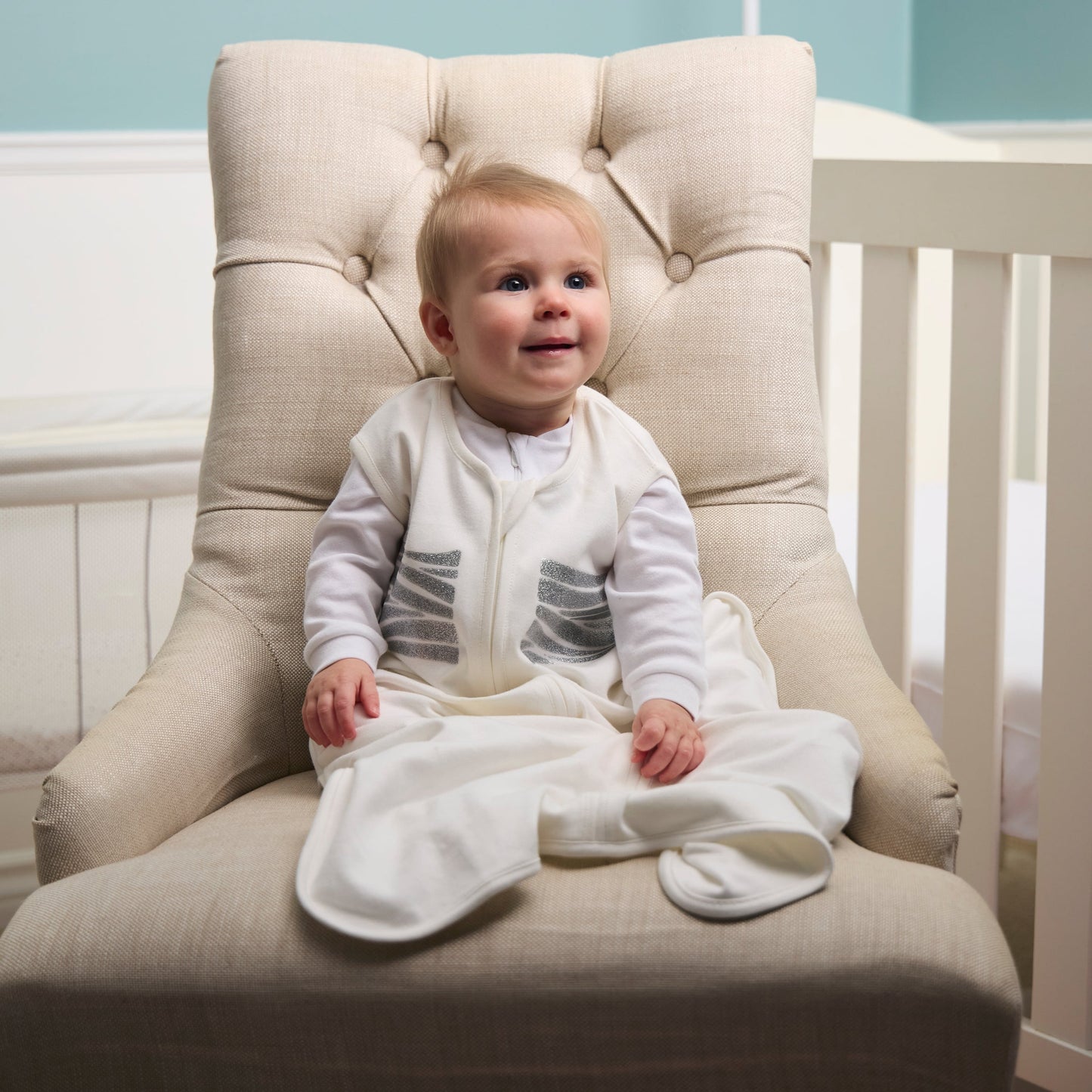 Baby in a white outfit sitting on a beige chair in a nursery.