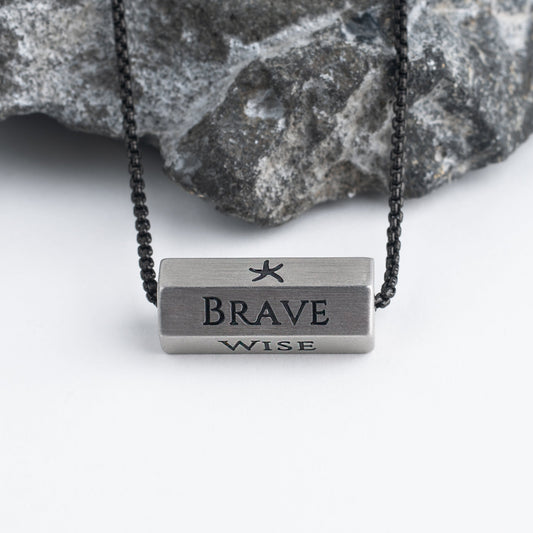 Pillar silver bar pendant engraved Brave and Wise on black chain against gray rock