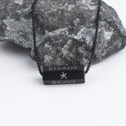 Pillar matte black bar pendant engraved Steady and Brave over gray stone
