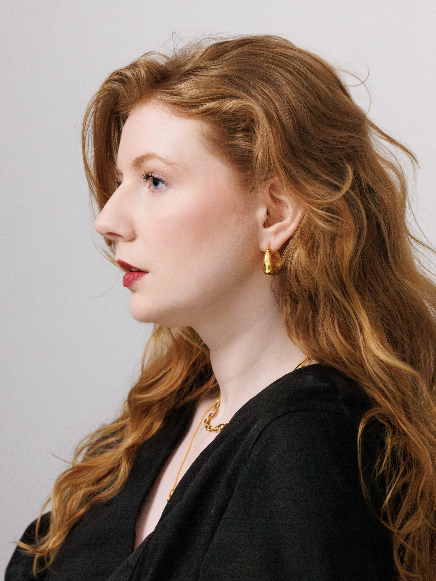 Woman with long, red, wavy hair wearing gold hoop earrings and a black top against a plain background