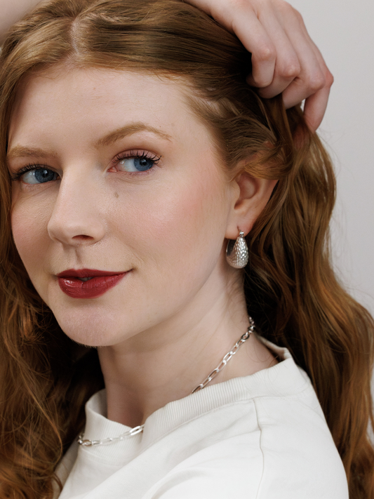 Woman with long red hair wearing silver hoop earrings and a silver chain necklace, with a neutral background
