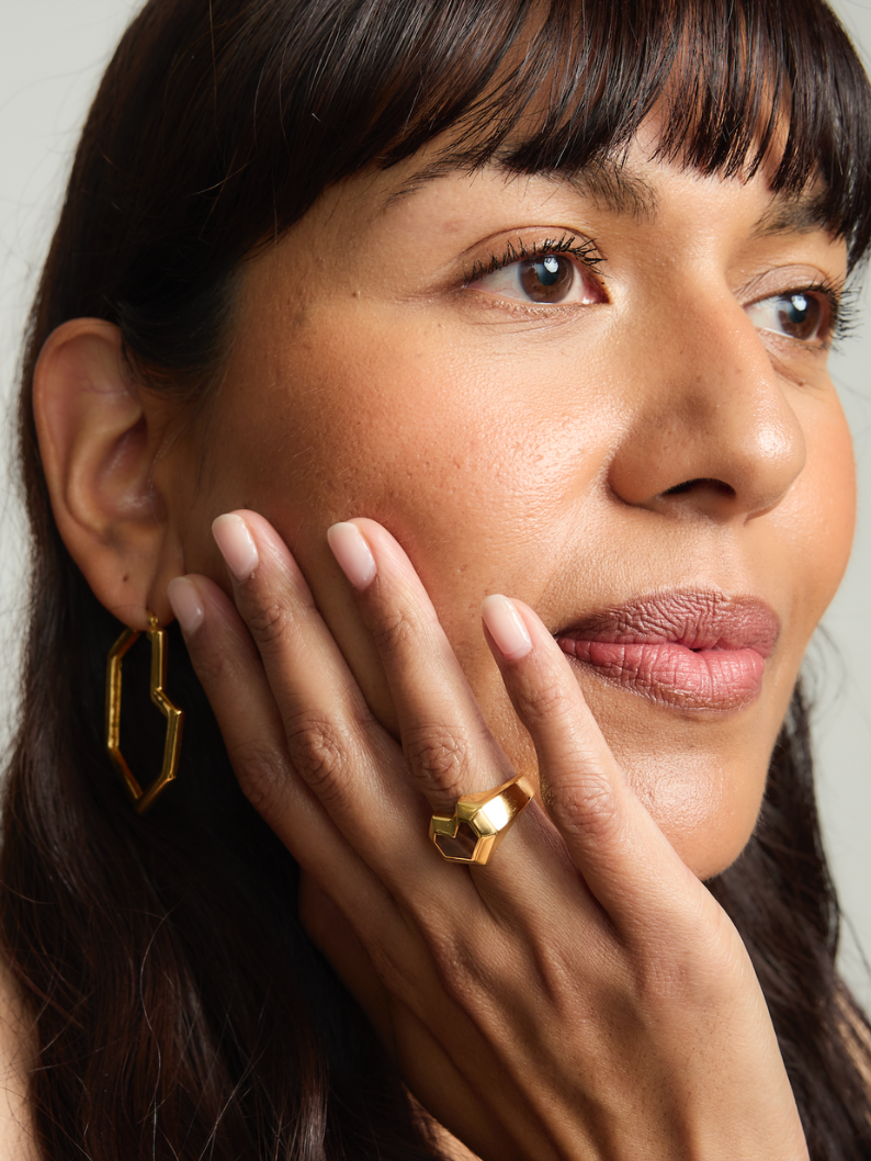 Close-up of a woman wearing gold hoop earrings and a ring, with a neutral background.