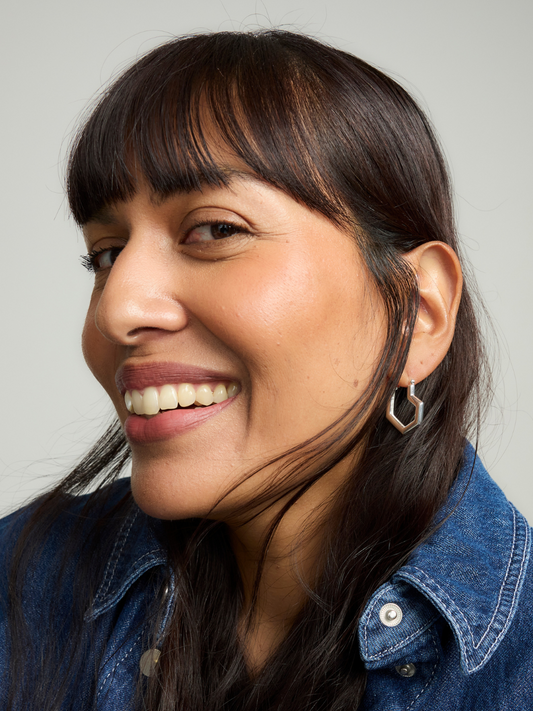 Woman with dark hair wearing silver heart hoop earrings smiling against a neutral background
