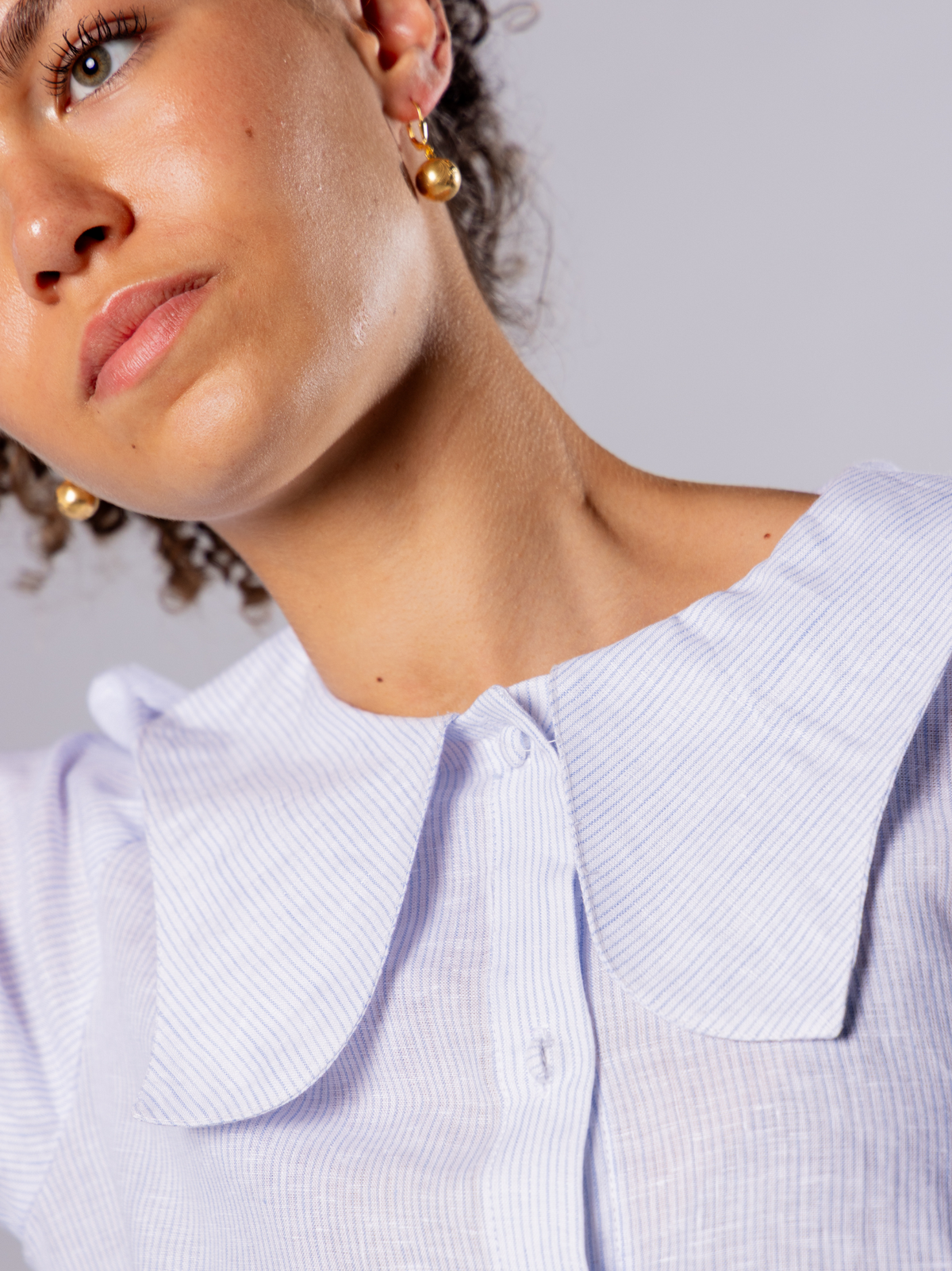 Close-up of a person wearing a light blue tulip collared shirt and gold drop ball earrings with a neutral background