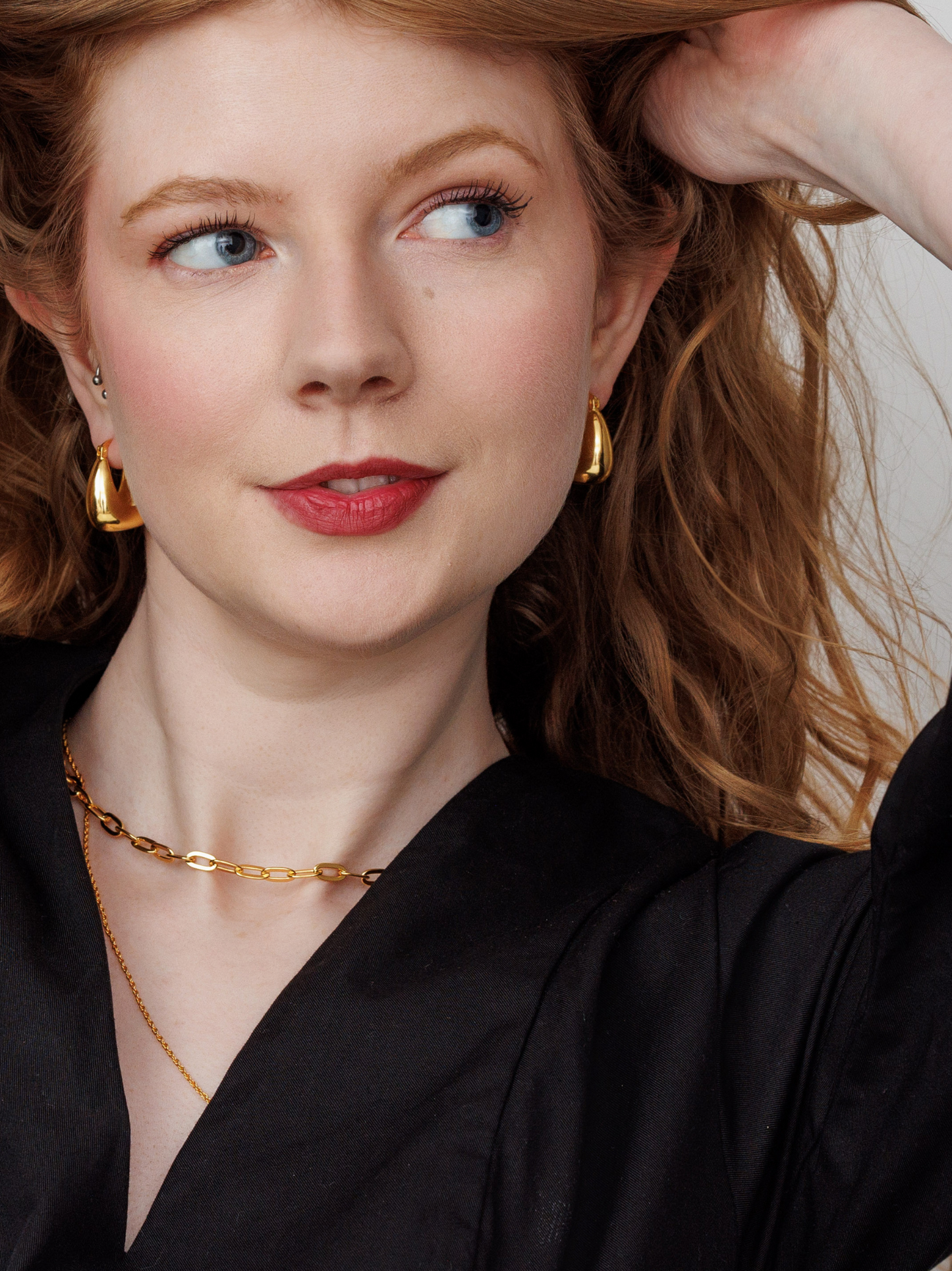 Woman with red hair wearing gold earrings and a chain necklace, against a neutral background