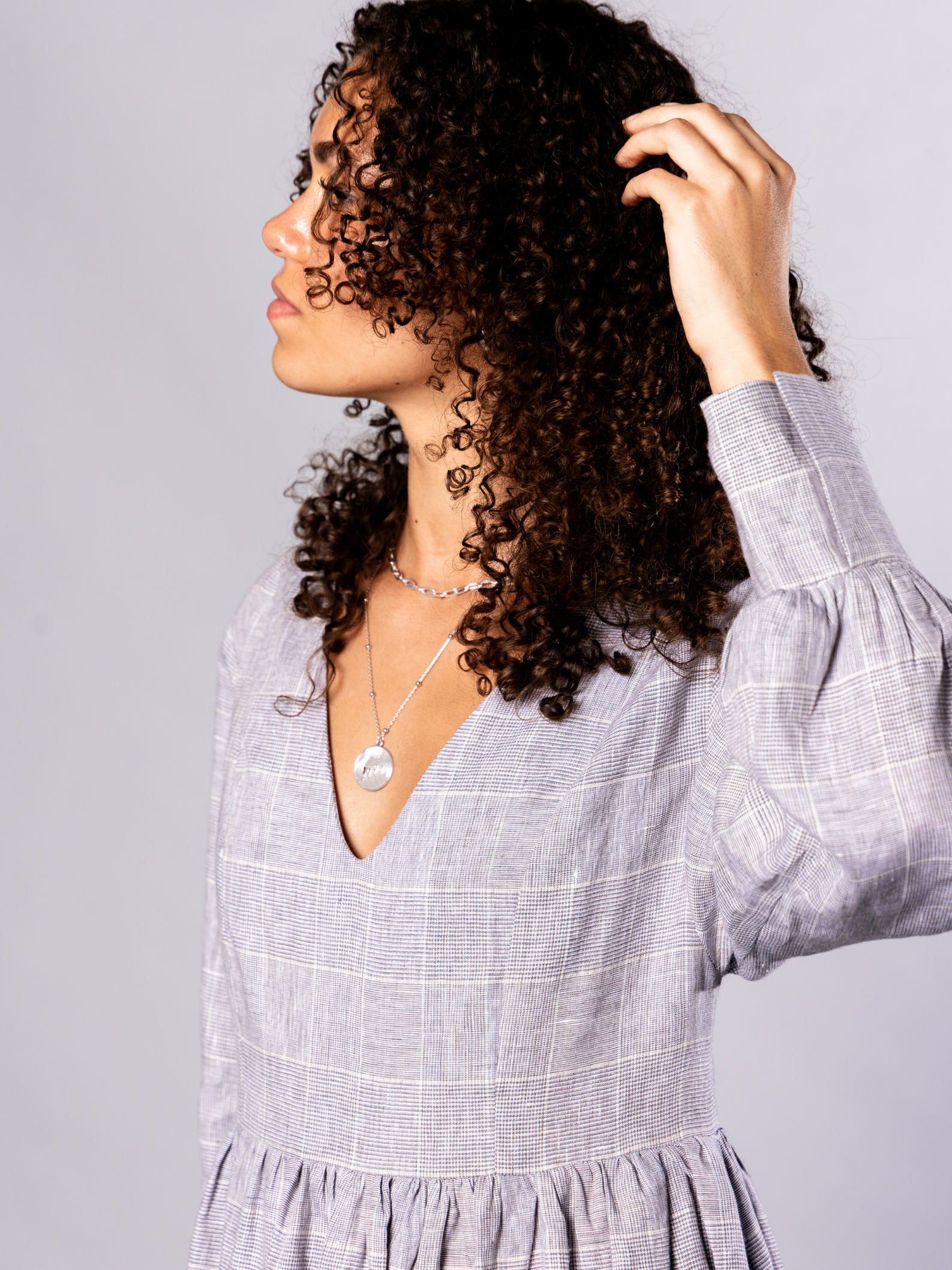 Woman wearing a light grey dress with silver chain and medallion necklace with a plain background