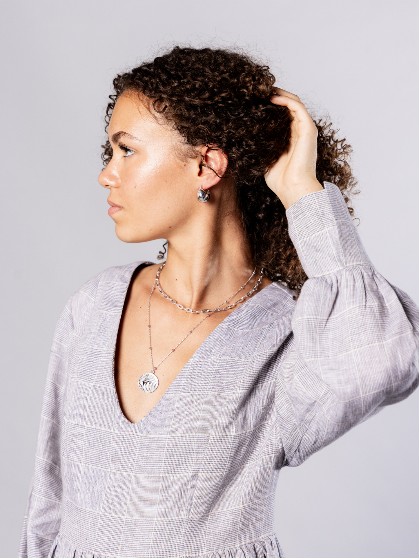 Woman adjusting her hair wearing recycled silver hoops and silver chain and medallion necklaces against a plain background