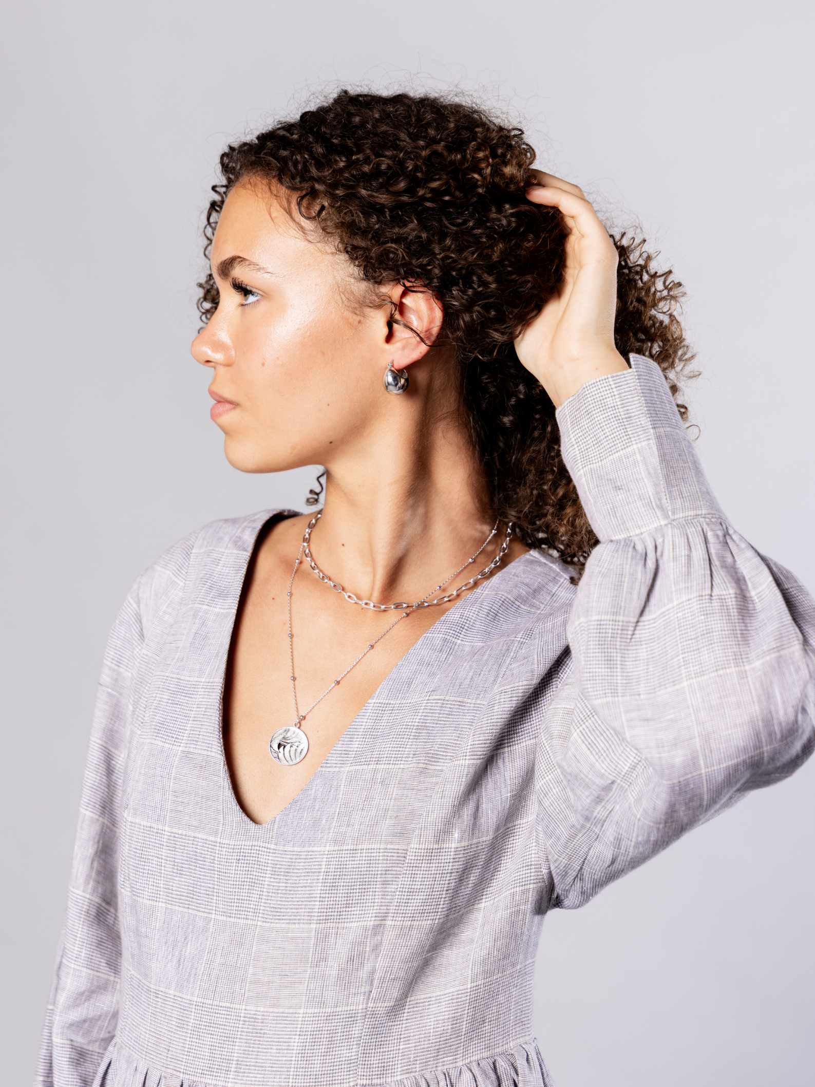 Woman adjusting her hair wearing recycled silver hoops and silver chain and medallion necklaces against a plain background