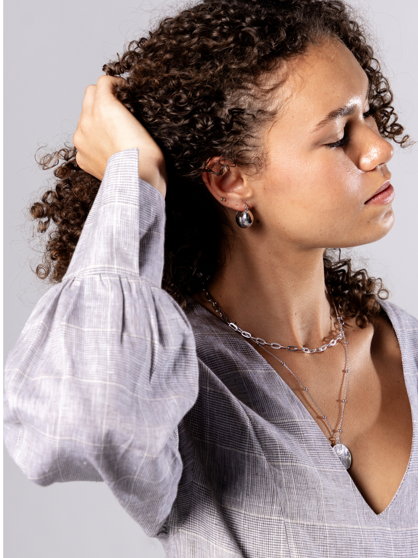 Woman wearing a light grey v neck dress with puffed sleeves and silver chain necklace  against a plain background