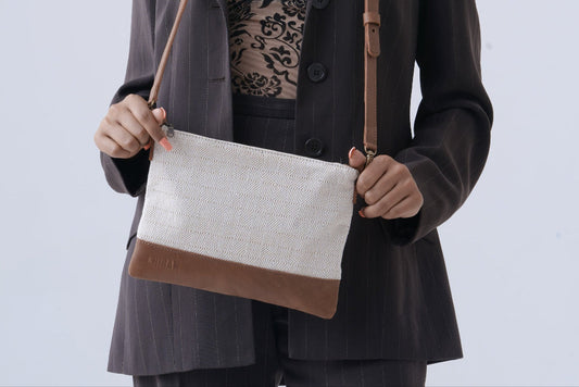 A model wearing a dark gray pinstripe blazer and black lace top holds a rectangular chiban bag with a white linen-textured body and brown leather trim and strap against a light gray studio background.