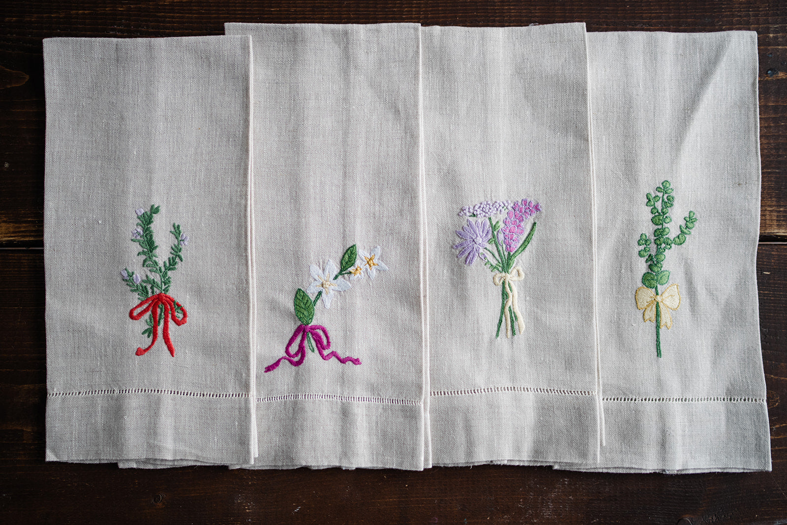 Folded flax linen tea towels with hand embroidered floral designs and a bow motif, displayed on a wooden surface.