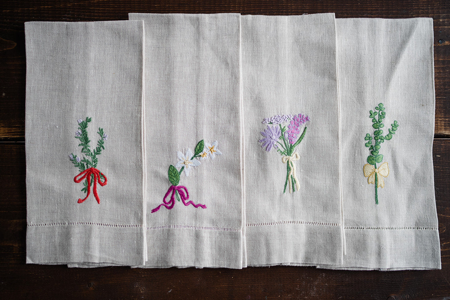 Folded flax linen tea towels with hand embroidered floral designs and a bow motif, displayed on a wooden surface.