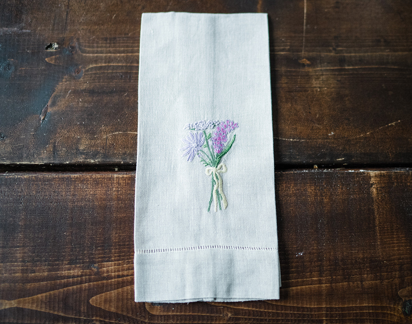 Folded flax linen tea towels with hand embroidered floral designs and a bow motif, displayed on a wooden surface.