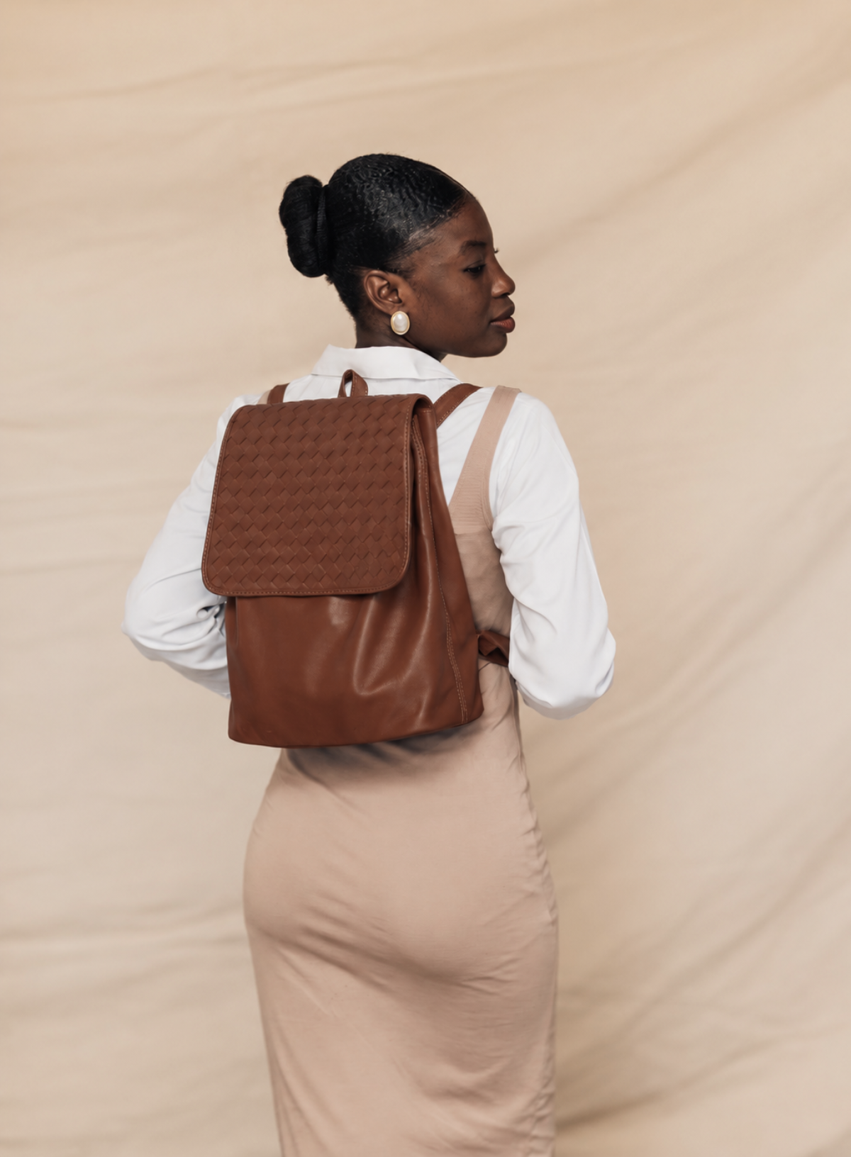 A woman wearing a brown leather backpack with a textured woven flap, styled with a white long-sleeve shirt and a beige dress against a beige fabric backdrop.
