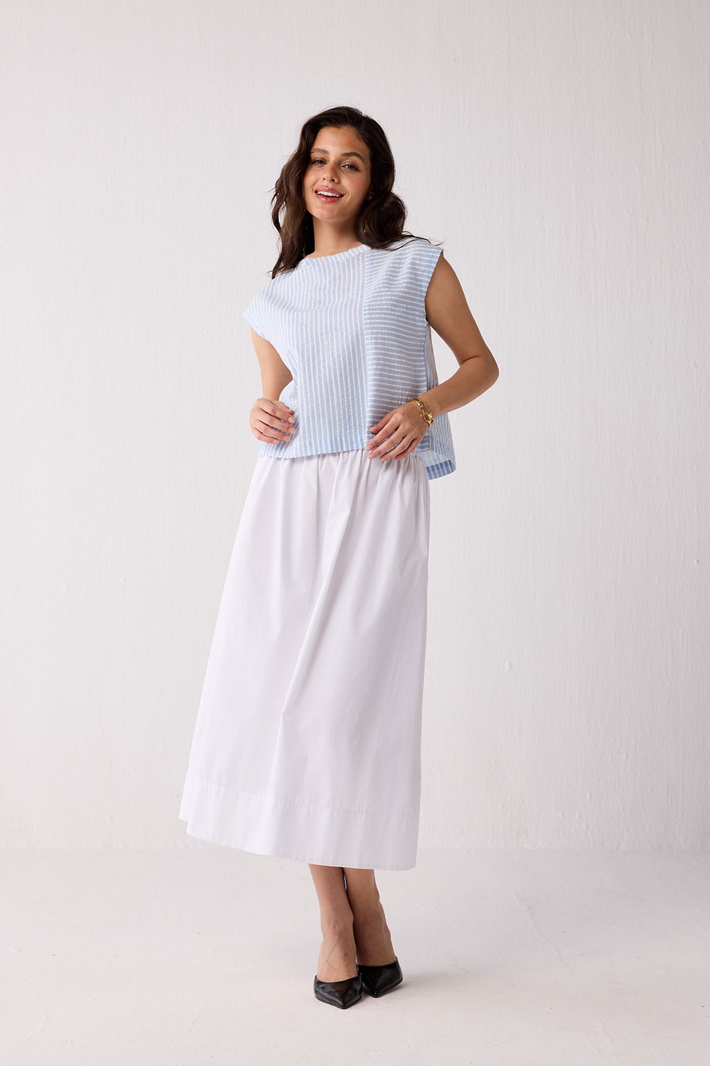 A woman wearing a light blue striped top and a white skirt on a plain background