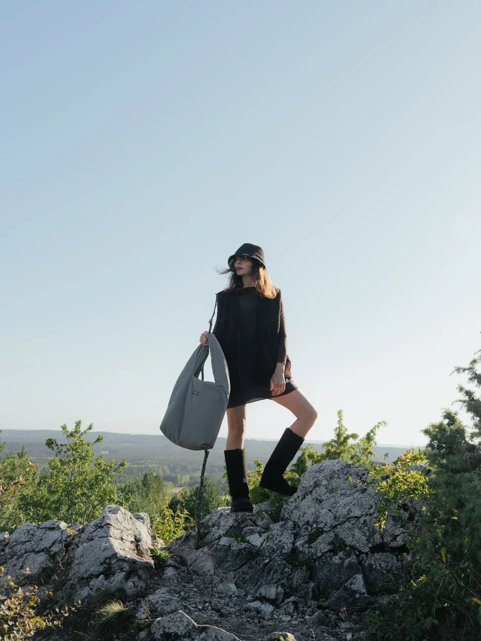 Person holding a gray bag on a rocky outcrop with a scenic background wearing long vegan boots