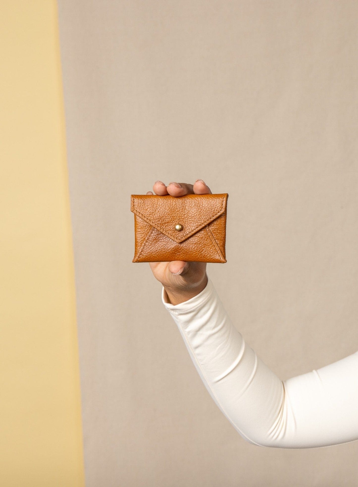 Person holding a cognac leather coin pouch against a beige background
