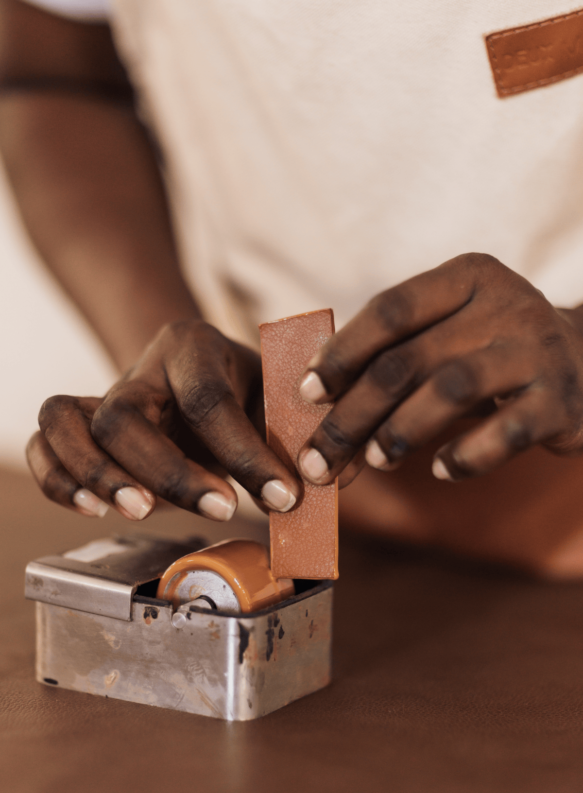 A close-up of a person's hands using a small metal leather roller tool on a tan leather strip, with a cream apron visible in the background.