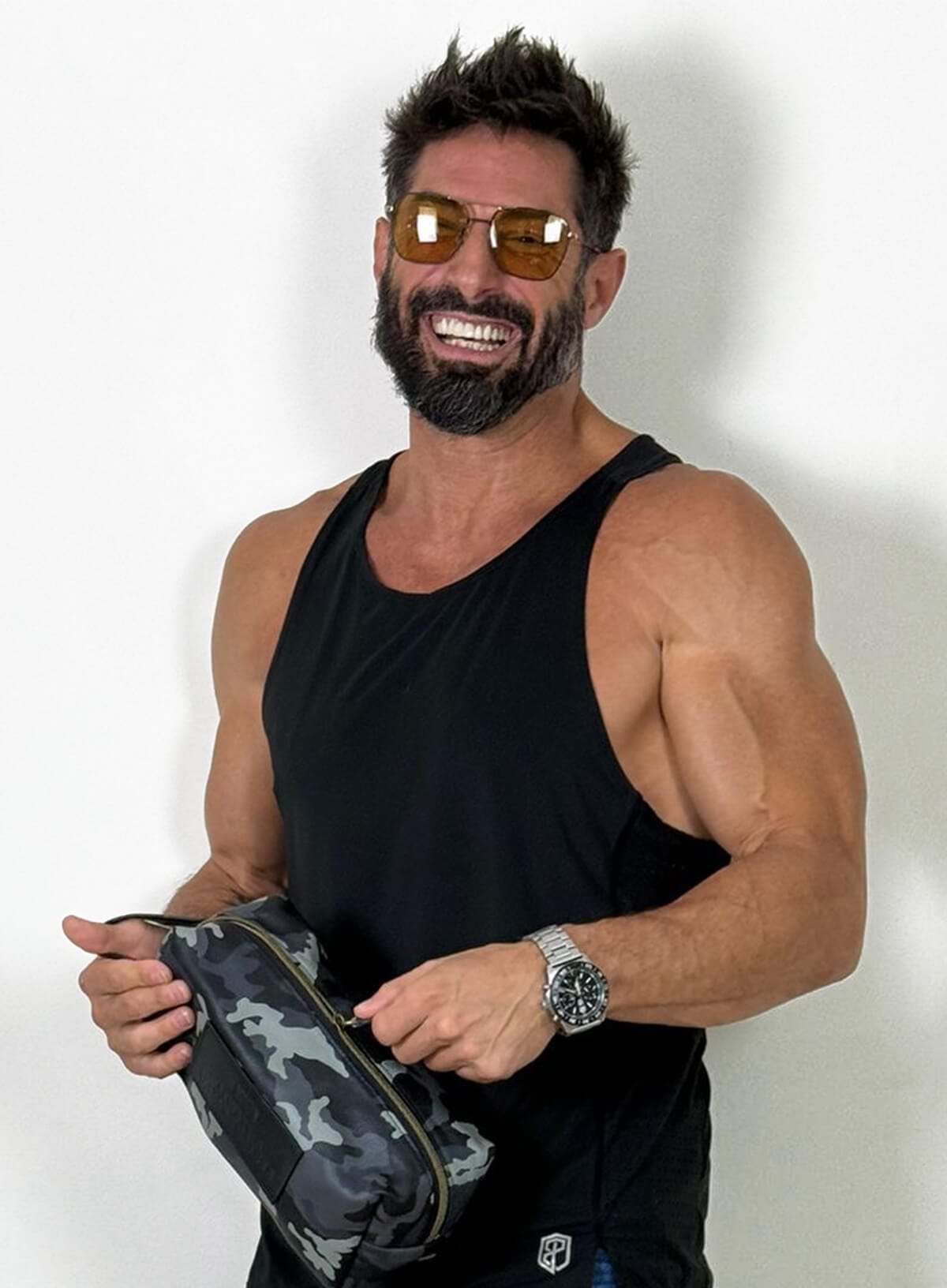 Man wearing sunglasses and a black tank top holding a camouflage bag against a white background
