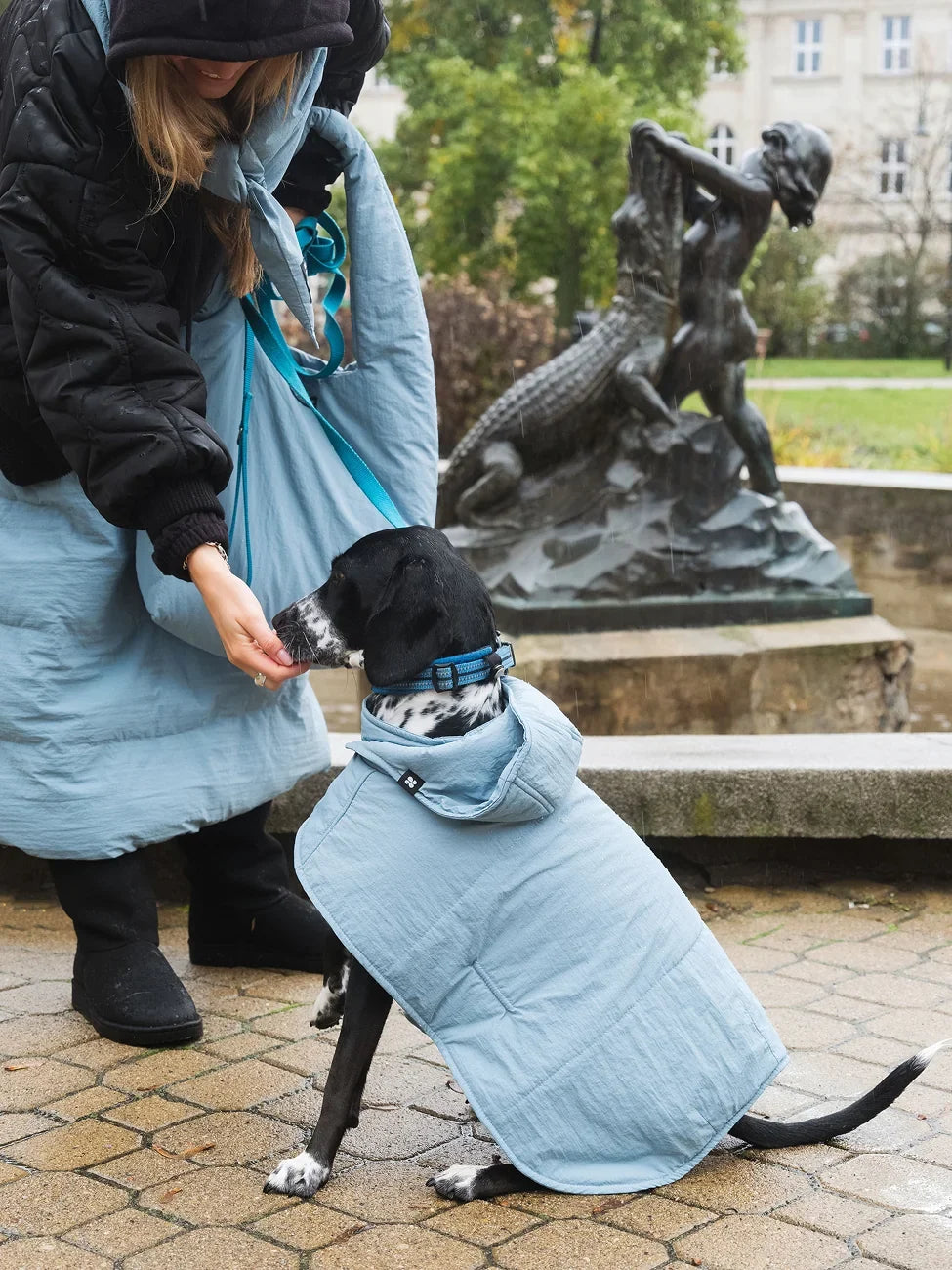 Person in a long light blue skirt with a dog wearing a matching coat, standing in an outdoor setting with a statue in the background.